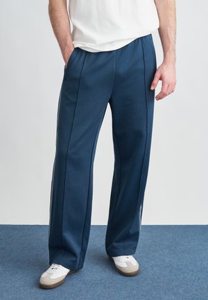 Marineblauwe ontspannen broek met plooien, zijzakken en contrasterende strepen aan de zijkant. Gemaakt van glad, rekbaar materiaal.