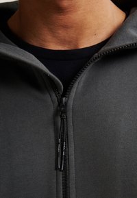 Sudadera gris con cremallera y textura suave, cremallera negra y detalle en la capucha. Llevada sobre una camiseta oscura, resaltando el estilo en capas.