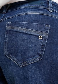 Jeans de mezclilla de un azul oscuro con un corte clásico, costuras visibles y un bolsillo trasero con un detalle metálico circular.