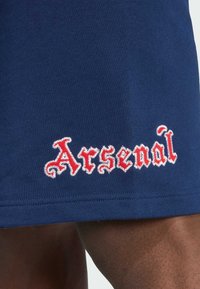 Mørkeblå shorts med ribbet tekstur; har hvid og rød broderet tekst "Arsenal" i en dekorativ skrifttype på venstre ben.