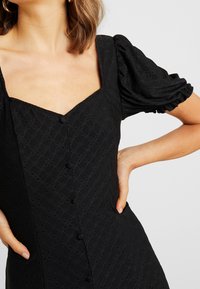 Svart klänning med puffärmar, fyrkantig uringning, texturerad tyg och dekorativa knappar fram. Tajt passform med slät yta.