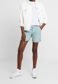 Henkilö, joka käyttää valkoista farkkutakkia, vaaleanharmaata t-paitaa, mintunvihreitä käärittyjä shortseja ja valkoisia korkearapaisen tennareita seisoen yksinkertaista taustaa vasten.