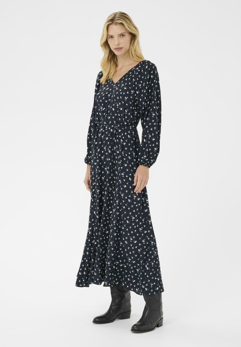 Langes marineblaues Kleid mit weißem Blumenmuster, V-Ausschnitt, langen Ärmeln und tailliertem Schnitt. Kombiniert mit schwarzen Stiefeln mit Absatz. Glatte Stoffstruktur.