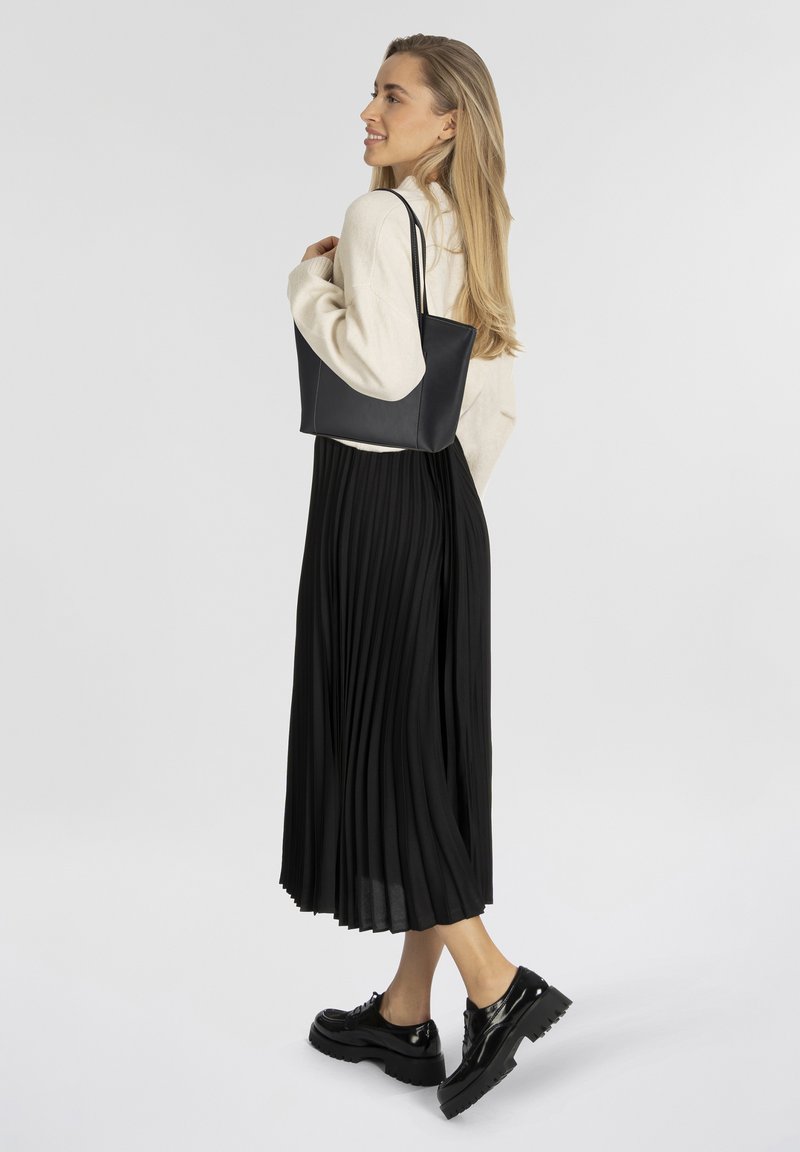Sac fourre-tout noir avec une texture en cuir lisse, présentant un design simple. Le modèle porte un haut crème et une jupe plissée noire, agrémentés de chaussures noires brillantes.