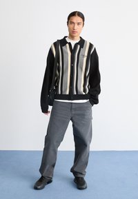 Cardigan lavorato a maglia con strisce verticali nere, crema e grigie, chiusura a bottoni frontale e maniche nere. Abbinato a pantaloni ampi grigi e scarpe nere.