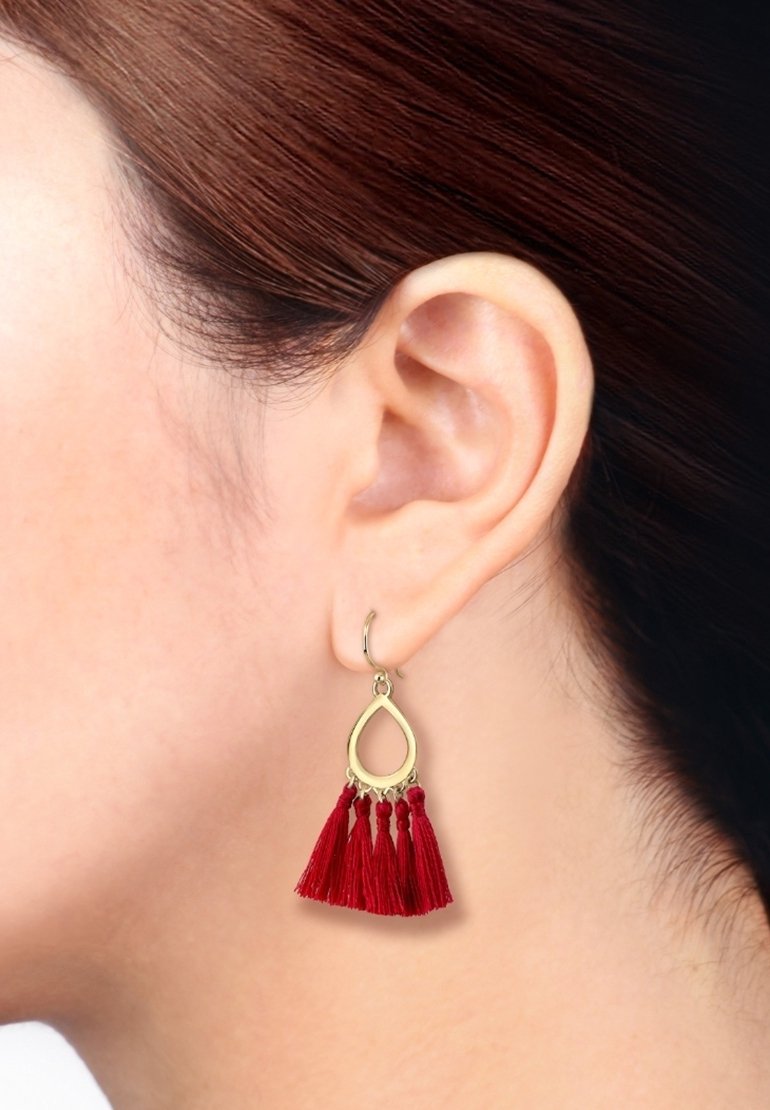 Gros plan sur l’oreille d’une femme portant une boucle d’oreille en forme de goutte dorée avec cinq pompons rouges suspendus.