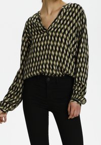 Femme portant un blouse à manches longues avec des rayures diagonales olive, noire et beige, rentrée dans un pantalon noir taille haute.
