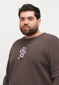 Brown sweatshirt featuring a vertical embroidered design of pink flowers on the front. The fabric appears soft with a relaxed fit.