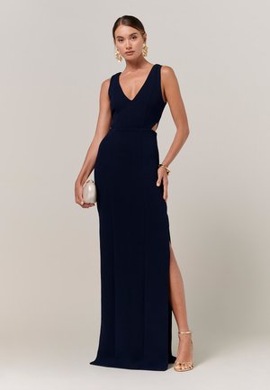 Frau trägt ein ärmelloses, marineblaues Abendkleid mit seitlichen Ausschnitten und hohem Schlitz, hält eine weiße Clutch und ist mit goldenen Ohrringen und High Heels ausgestattet.