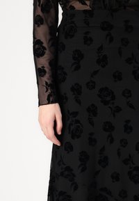 Μαύρο φόρεμα με λουλουδάτο σχέδιο και διάφανα μακριά μανίκια. Το ύφασμα έχει απαλή υφή, με ανάγλυφα μαύρα τριαντάφυλλα σε ματ φόντο.