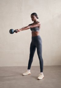 Schwarze Sportleggings mit einem strukturierten Muster, einem passenden grauen Sport-BH und beigen Turnschuhen. Die Person hält ein blaues Kettlebell.