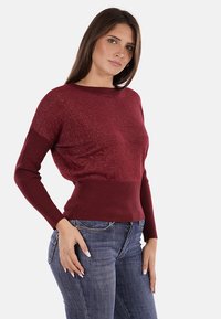 Maglione in maglia cremisi con cintura e polsini a coste, caratterizzato da un corpo morbido e texture e maniche lunghe. Silhouette aderente con collo rotondo.