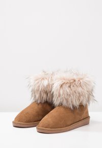 Botas de ante marrón con un puño de piel de oveja beige y esponjoso, diseño de punta redonda y suela de goma plana. Textura suave con una apariencia acogedora.