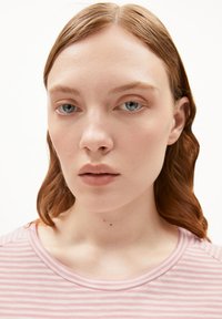 Modèle portant un haut rayé rose clair avec un col rond. Les cheveux sont ondulés et de longueur moyenne, et la peau semble lisse avec un maquillage minimal.