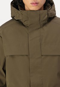 Veste imperméable vert olive avec un col haut, des lignes épurées, des coutures minimales et une texture lisse. Présente un rabat avant dissimulé.