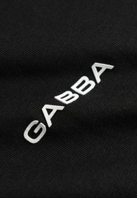 Logo texte blanc GABBA sur tissu noir texturé, légèrement incliné et surélevé par rapport à la surface.