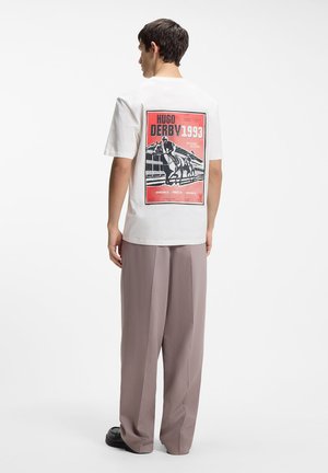 Man wearing a white t-shirt with a red and black horse racing graphic and "Hugo Derby 1993" text on the back, paired with loose beige pants.