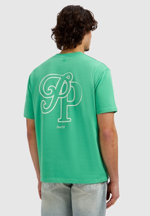 Man with curly dark hair wearing a green t-shirt with large white "PP" letters and "PurePath" text on the back, paired with light denim jeans.
