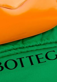 Sac à main en cuir orange sur tissu vert avec une partie du mot "BOTTEGA" imprimée en noir.