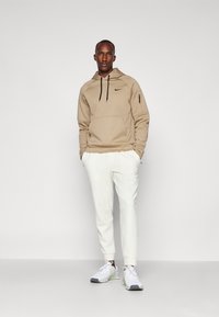 Beige hoodie with a kangaroo pocket and black drawstrings, paired with white tapered trousers and white athletic shoes with gray accents.