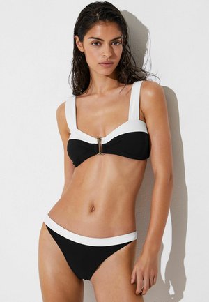 Kobieta z mokrymi włosami, ubrana w czarne bikini z szerokimi białymi paskami i wykończeniem, stojąca na tle białej ściany z cieniami rzucanymi za nią.