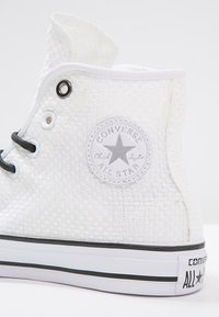 Bílé tkané plátěné tenisky Converse Chuck Taylor All Star vysoké s černými tkaničkami a logem na kotníku.