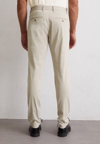 Beige katoenen broek met elastische tailleband, voorzien van twee achterzakken met knoopsluitingen en een rechte pijpuitvoering. Gekozen in combinatie met zwarte schoenen.