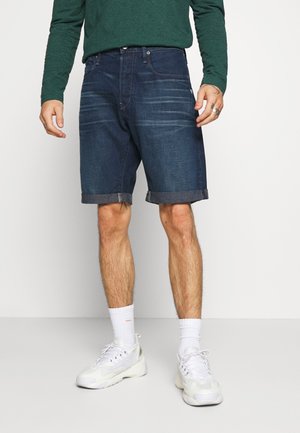 Homme portant un short en denim bleu foncé retroussé, une chemise verte à manches longues, des chaussettes blanches et des baskets blanches sur un fond uni.