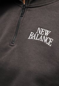 Πουλόβερ σε σκούρο γκρι ύφασμα, με εμπρός φερμουάρ μέχρι τη μέση και λευκό κεντημένο λογότυπο "NEW BALANCE". Μαλακή υφή με casual σχεδιασμό.