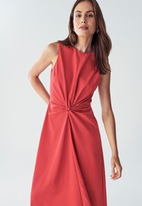 Červené bezrukávové šaty s prekrúteným detailom vpredu, s prúdiacou látkou a uvoľneným strihom, s dĺžkou po kolená a hladkou textúrou.