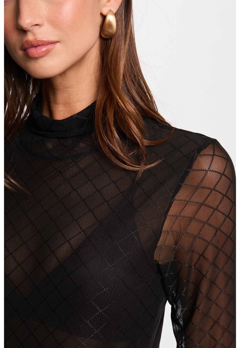 Femme portant un haut en mesh noir transparent avec un motif en diamond et de grandes boucles d'oreilles rondes en or, cheveux bruns sur l'épaule.