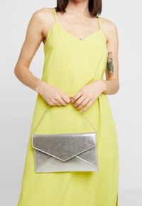 Pochette métallique argentée au design enveloppe, avec une surface texturée et une lanière chaîne, tenue par une personne portant une robe jaune.