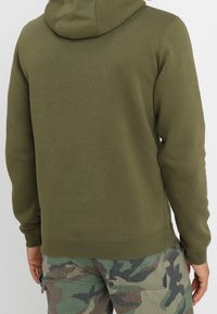 Oliivinvihreä huppari pehmeällä tekstuurilla, jossa on kengurutasku ja kiristysnauhallinen huppu. Yhdistetty maastokuvioisiin cargo-housuihin.