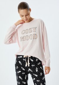 Vaaleanpunainen pitkähihainen paita, jossa on "COZY MOOD" -grafiikka, yhdistettynä mustiin pyjamahousuihin, joissa on valkoisia jääkarhuja ja tähdistökuvio.