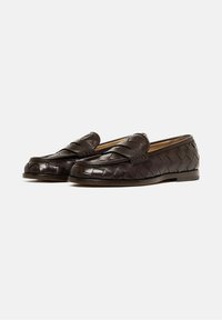 Par de mocasines de cuero trenzado marrón oscuro con tira tipo penny, tacón bajo y punta redondeada sobre un fondo blanco.