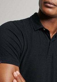 Czarny bawełniany t-shirt polo z kołnierzykiem, krótkimi rękawami oraz małą metką marki na rękawie. Gładka tekstura i dopasowany krój.