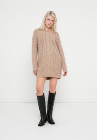 Beige knitted sweater dress with cable-knit texture, long sleeves, and ribbed hem, paired with black knee-high boots.
