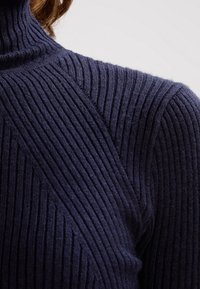 Pull col roulé en tricot côtelé bleu marine avec une surface texturée, une coupe ajustée, et des coutures diagonales au niveau de l'épaule.