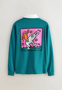 Tyrkysová dlouhá rukávová košile s velkým růžovým potiskem na zádech, zobrazující stylizovanou postavu, jak jezdí na rybě proti modrým vlnám.