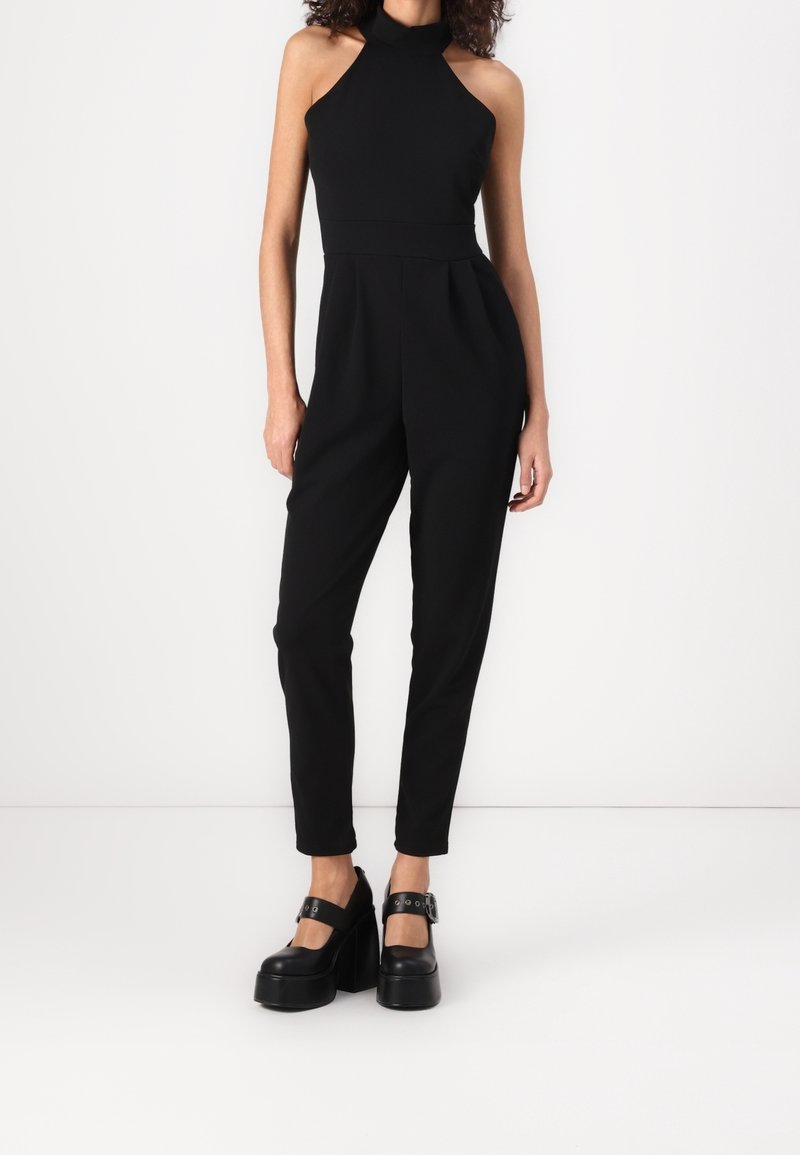 Femme portant une combinaison noire sans manches à col halter et des chaussures Mary Jane noires à plateforme, debout devant un fond blanc.