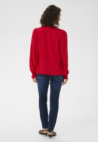 Blouse rouge à manches longues avec épaules bouffantes, poignets ajustés et col montant, associée à un jean slim bleu foncé et des sandales noires.