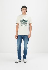 Tricou din bumbac bej, cu un imprimeu grafic verde și textul "GRAND PALACE HOTEL". Asortat cu blugi albaștri și adidași albi cu accente negre.