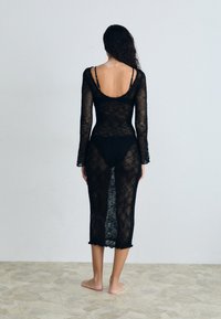 Vestito nero a maniche lunghe in tessuto a rete, con apertura sul retro e bordo a scallop, trasparente in tutto, sovrapposto a un bikini nero.