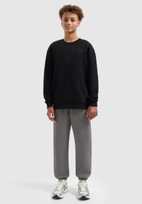 Zwarte crewneck sweatshirt met lange mouwen, gecombineerd met grijze joggingbroek. Model draagt lichtgekleurde sneakers. Soepele stof, relaxte pasvorm.