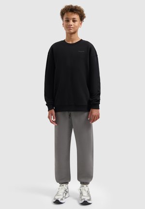 Zwarte crewneck sweatshirt met lange mouwen, gecombineerd met grijze joggingbroek. Model draagt lichtgekleurde sneakers. Soepele stof, relaxte pasvorm.