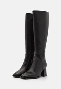 Bottes hautes en cuir noir pour femmes avec talons carrés et fermetures éclair latérales, présentées debout sur un fond blanc.
