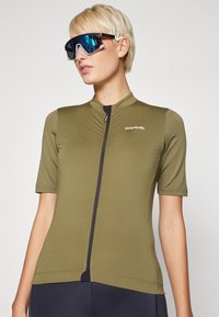 Jersey de ciclismo de manga corta en tejido verde oliva, con cremallera frontal y costuras planas. Combinado con unas grandes gafas de sol reflectantes.