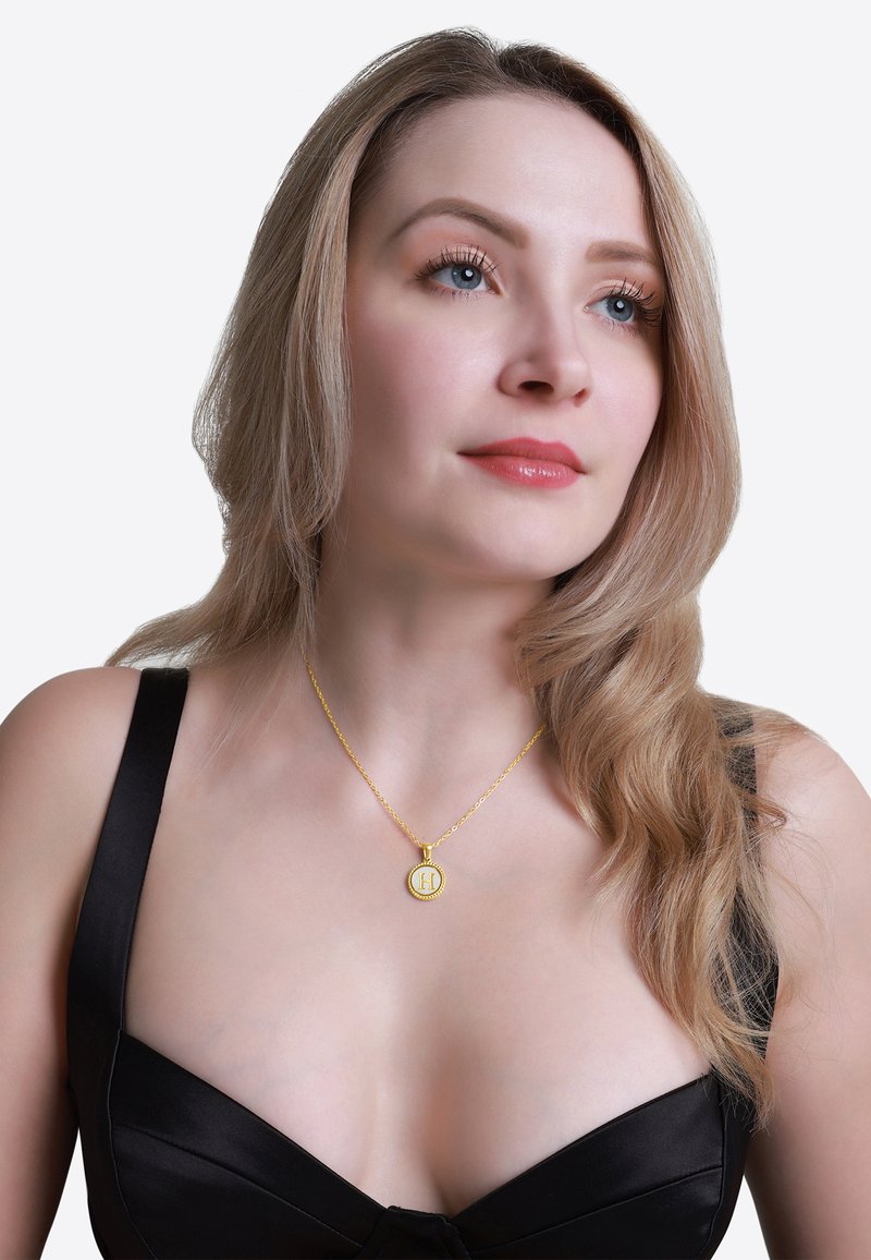 Złota naszyjnik z wisiorkiem w okrągłym kształcie z ozdobnym brzegiem, z literą "H", zawieszony na łańcuszku noszonym na czarnej bluzce.