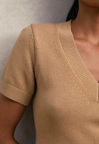 Világosbarna, rövid ujjú, V nyakú kötött pulóver. A nyakrészen és az ujjaknál texturált bordázat található, amelyen sima anyagfelület figyelhető meg.
