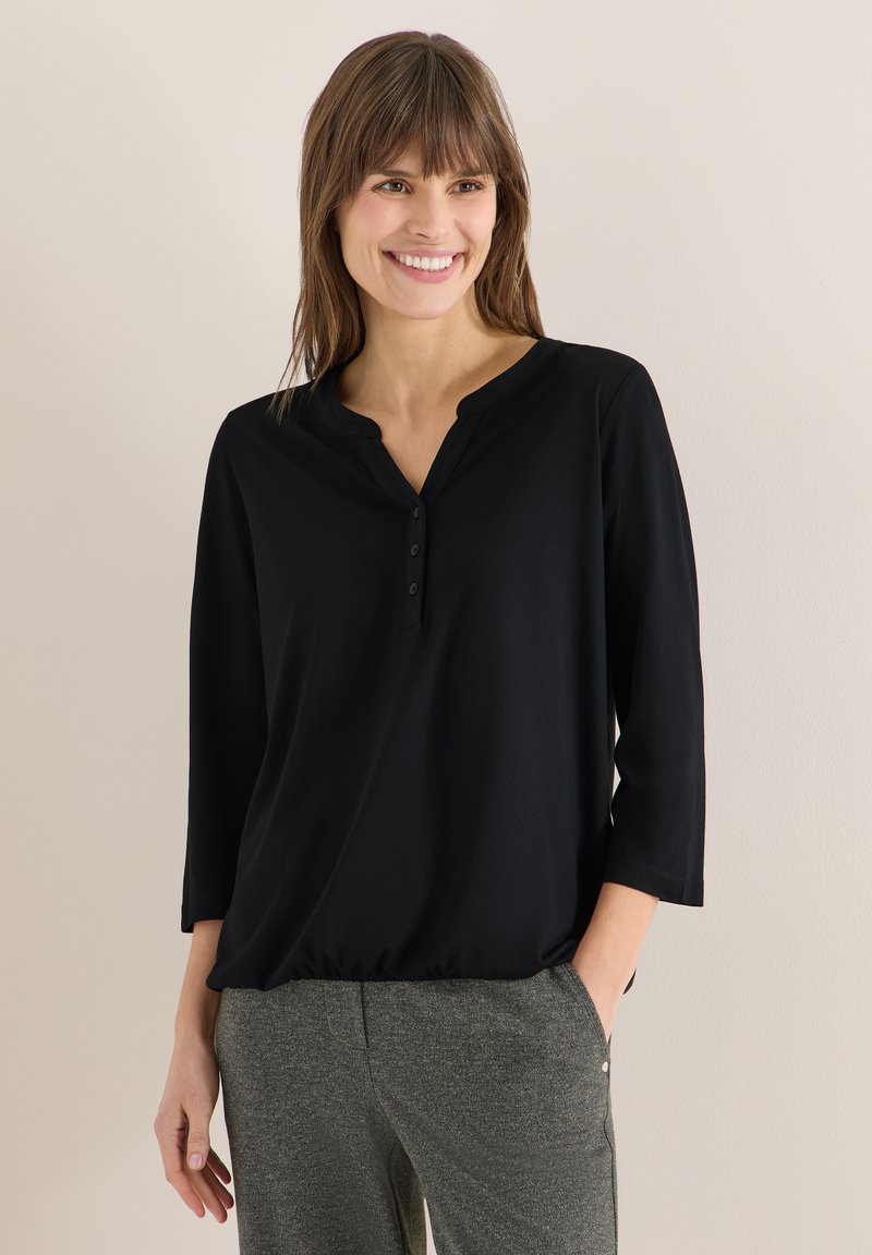 Femme souriante aux cheveux bruns portant un haut henley noir à manches longues et un pantalon gris, debout avec une main dans la poche.